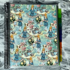 May include: A blue and white patterned spiral bound notebook with a tea party theme. The cover features illustrations of teacups, teapots, and girls in dresses. The text on the cover reads "Time for Tea Party" and "You're Invited".