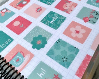 May include: A white planner with a clear plastic cover. The cover has a grid pattern and is decorated with colorful stickers featuring floral designs, words like "hello" and "hi!" and other illustrations. The planner has a black coil binding.
