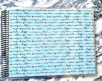 May include: A blue planner cover with a clear plastic front. The cover has a repeating pattern of black cursive text that says "imagine", "believe", "magic", "say cheese", "mouse & co", "dream", "smile", "happy" and red hearts.