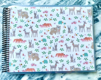 May include: A white spiral bound notebook with a clear plastic cover. The cover features a repeating pattern of woodland animals, including deer, foxes, bears, and rabbits. The animals are illustrated in a cartoon style and are surrounded by leaves and flowers.
