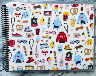 May include: A white planner with a colorful pattern of various objects, including cameras, ice cream cones, pretzels, sunglasses, and backpacks. The planner has a clear plastic cover and a black spiral binding.