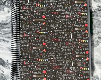 May include: A spiral-bound notebook with a black cover featuring a school-themed pattern. The design includes illustrations of pencils, apples, books, and text such as "School Days," "Math," and "Learning." The notebook is encased in a clear plastic cover.
