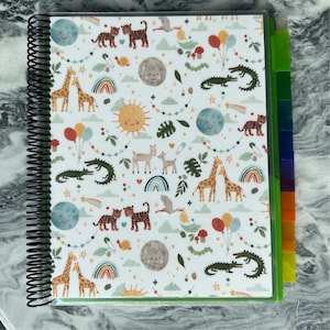 May include: A white spiral bound notebook with a green cover and colorful illustrations of animals, stars, rainbows, and clouds. The notebook has multiple colored tabs on the side.
