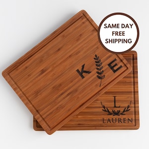 May include: Two wooden cutting boards with engraved initials and a leafy design. The top board has the initial "K" and the bottom board has the initial "L" with the name "Lauren" underneath.