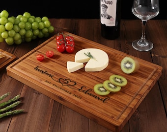 Puede incluir: Una tabla de cortar de bambú personalizada con los nombres "Kristene" y "Edward" grabados en ella, junto con la fecha "EST. DICIEMBRE 2021". La tabla está decorada con un diseño de corazón y presenta una cuña de queso, rodajas de kiwi y tomates cherry.