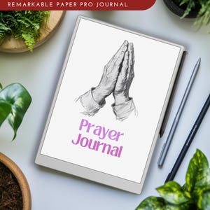 Puede incluir: Un bloc de notas digital blanco con un dibujo de manos juntas en oración. El texto "Prayer Journal" está escrito en rosa en la pantalla. El bloc de notas está sobre una superficie blanca con un lápiz táctil negro y plateado.