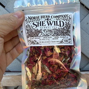 May include: A holographic resealable pouch containing loose leaf herbal tea. The white label reads Norse Herb Company She Wild Herbal Tea, listing organic peppermint, ginger pieces, organic raspberry leaf, red rose petals, and mugwort as ingredients.