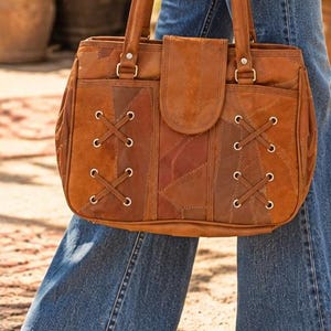 May include: A brown leather handbag featuring a patchwork design with visible stitching. The front panel includes two decorative lace-up sections with silver-toned metal grommets and a central flap closure. It has two sturdy top handles.