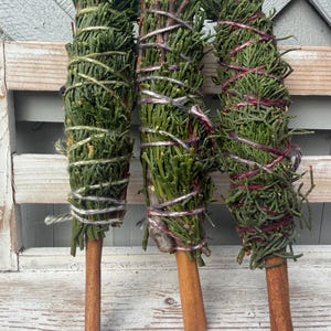 May include: Three bundles of green cedar needles wrapped with colored twine and attached to a brown cinnamon stick handle. The bundles are arranged vertically, each featuring a distinct color of string: light green, purple, and dark red.