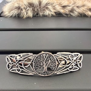 May include: A silver-toned metal hair barrette featuring a central circular medallion with a detailed tree design. The barrette is flanked on both sides by intricate, symmetrical Celtic knotwork patterns, creating a cohesive and ornate hair accessory.