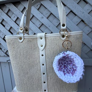 May include: A beige tote bag with cream-colored handles and trim. The bag has a textured fabric body and a vertical cream-colored strap with rivets. A decorative keychain with a purple and white geode design hangs from the bag.