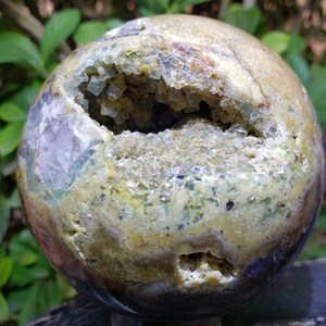 May include: A spherical geode with a large opening revealing a cluster of sparkling crystals. The exterior features a mix of earthy tones, including brown, green, and hints of purple. The geode is a natural, decorative item.
