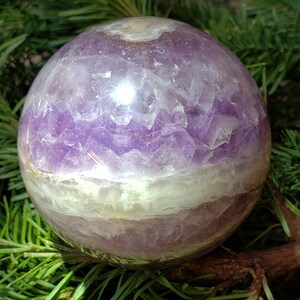 May include: A polished amethyst sphere, showcasing bands of purple and white. The crystal ball is resting on a bed of green pine needles and branches, creating a natural contrast. The sphere's surface reflects light, highlighting its smooth texture.
