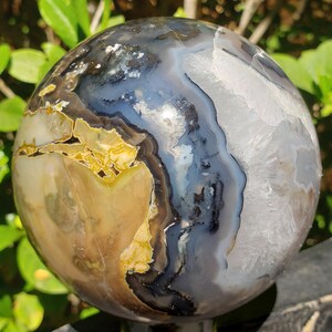 May include: A polished agate sphere with a mix of colors including beige, gray, black, and white. The sphere has a smooth, rounded shape and displays natural patterns and textures. This decorative stone is a unique home decor item.