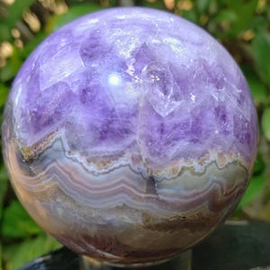 May include: A polished amethyst sphere, showcasing a blend of deep purple and lighter lavender hues. The lower portion reveals banded layers of white, tan, and grey, creating a natural, earthy aesthetic. The sphere is mounted on a silver-colored stand.