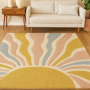 Puede incluir: Una alfombra rectangular con un diseño de sol. La alfombra presenta un sol amarillo en la parte inferior, con rayos ondulados en colores pastel de rosa, azul y amarillo. La alfombra está colocada sobre un suelo de madera.