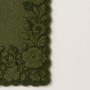 May include: A dark olive green placemat with a scalloped edge. The placemat features a detailed floral design in a raised, textured pattern. The flowers and leaves are in the same shade of green as the background.