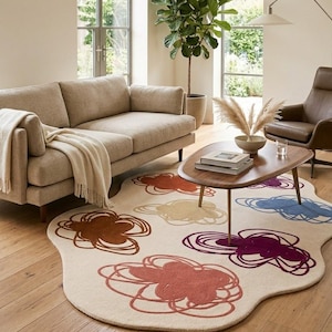 Puede incluir: Un salón con un sofá beige, un sillón de cuero marrón y una alfombra única. La alfombra tiene un fondo blanquecino con diseños florales coloridos en tonos rojos, marrones, morados y azules. Una mesa de centro de madera y una planta en maceta completan la decoración.