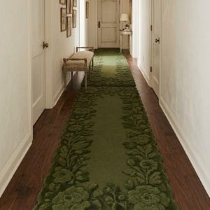 May include: A long, green floral runner rug with a scalloped edge, placed on a dark wood floor in a hallway. The rug features a raised floral design and adds a touch of elegance to the space.