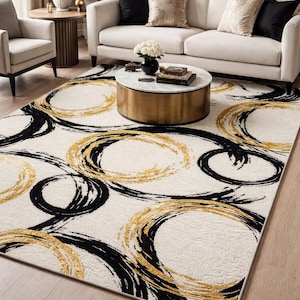 May include: A cream-colored area rug with a bold design of overlapping black and gold brushstroke circles. The rug is rectangular and placed on a hardwood floor, with a sofa and coffee table in the background.