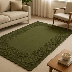 May include: A rectangular olive green rug with a floral border. The rug is placed on a light beige carpet in a living room setting. A beige sofa and armchair are visible in the background, along with a wooden coffee table.