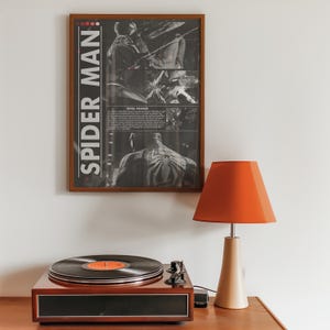 Op de afbeelding: Een ingelijste "Spider-Man" poster hangt boven een platenspeler en een tafellamp. De poster toont zwart-wit stripboekpanelen van Spider-Man. De platenspeler is bruin met een zwarte plaat. De lamp heeft een beige voet en een oranje kap.