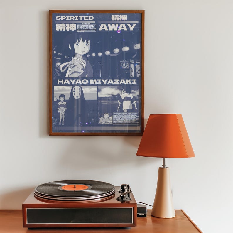 May include: A framed poster with a blue background featuring a character from the animated film Spirited Away. The poster has the title "Spirited Away" in Japanese and English, and the name "Hayao Miyazaki" in white text. The poster is hanging on a white wall above a record player and a lamp.