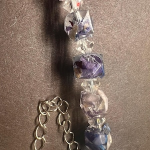 May include: A close-up of a necklace featuring clear, faceted beads and square resin beads with dried flowers in shades of purple, red, and white. The necklace has a silver chain.