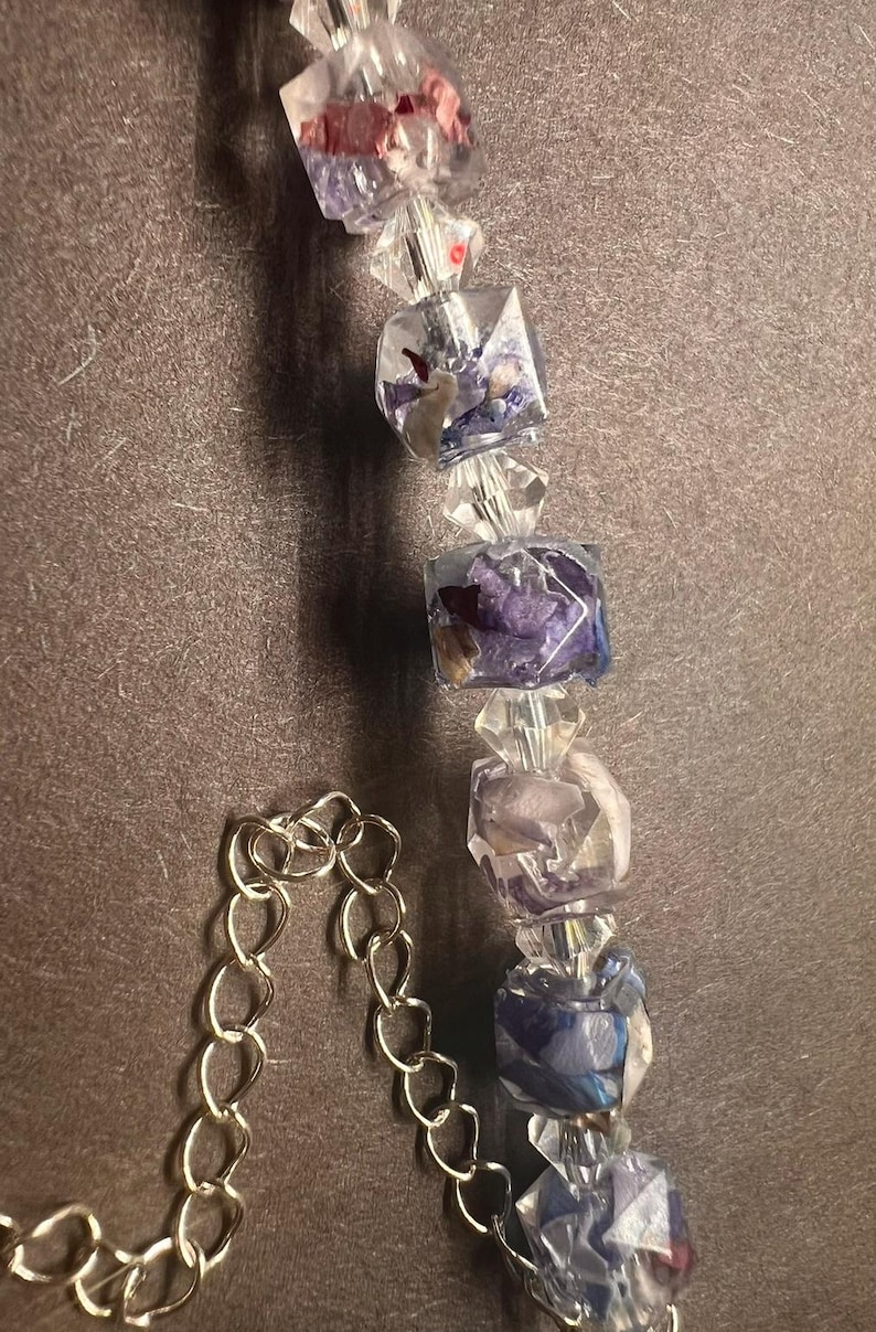 May include: A close-up of a necklace featuring clear, faceted beads and square resin beads with dried flowers in shades of purple, red, and white. The necklace has a silver chain.