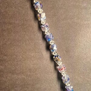 May include: A delicate bracelet featuring a series of colorful, square-shaped beads interspersed with clear, faceted beads. The beads display shades of blue, pink, and purple. The bracelet is finished with a silver-toned chain and a small cross charm.