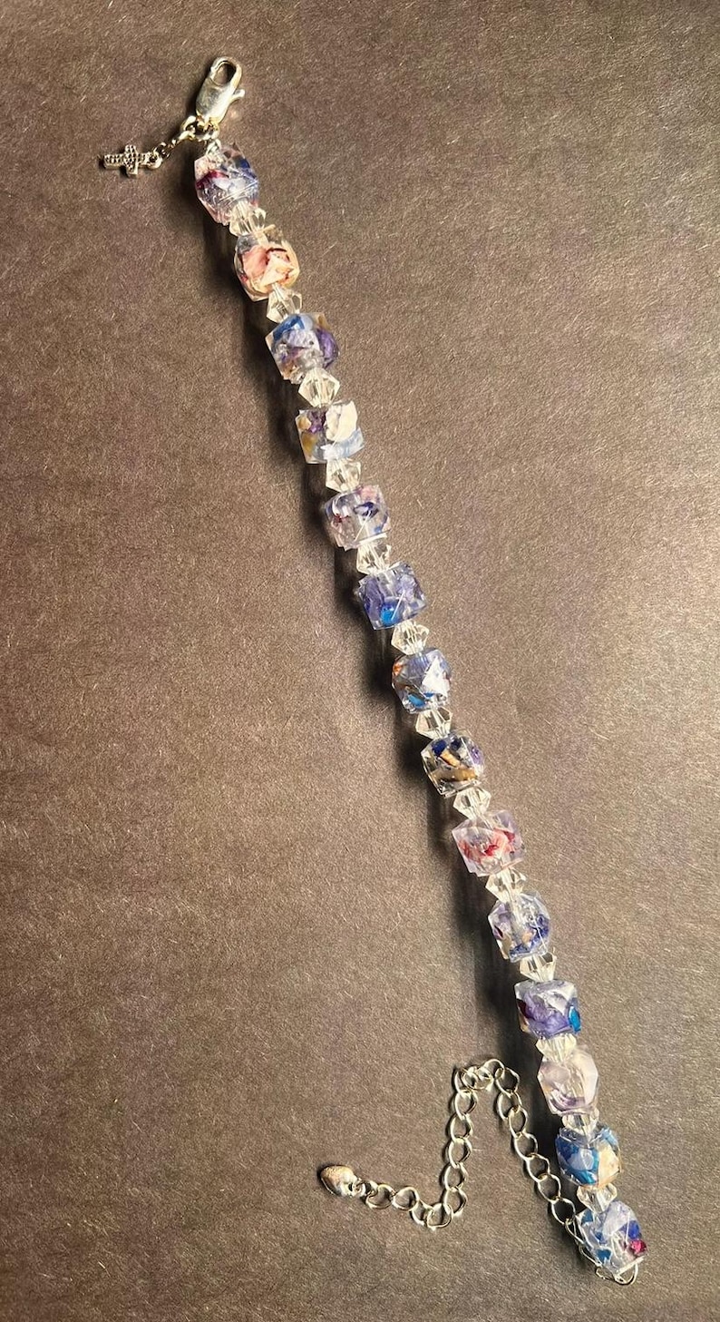 May include: A delicate bracelet featuring a series of colorful, square-shaped beads interspersed with clear, faceted beads. The beads display shades of blue, pink, and purple. The bracelet is finished with a silver-toned chain and a small cross charm.