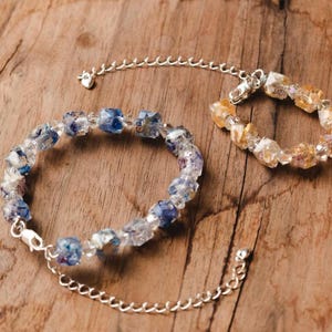 May include: Two beaded bracelets with silver-tone chain extenders rest on a wooden surface. One bracelet features blue and clear beads, while the other has yellow and clear beads. The bracelets are likely for jewelry.