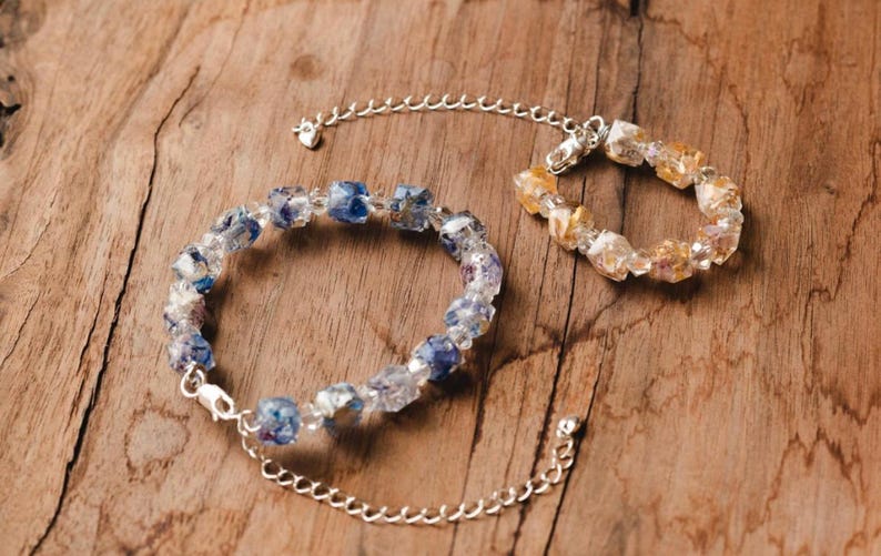 May include: Two beaded bracelets with silver-tone chain extenders rest on a wooden surface. One bracelet features blue and clear beads, while the other has yellow and clear beads. The bracelets are likely for jewelry.