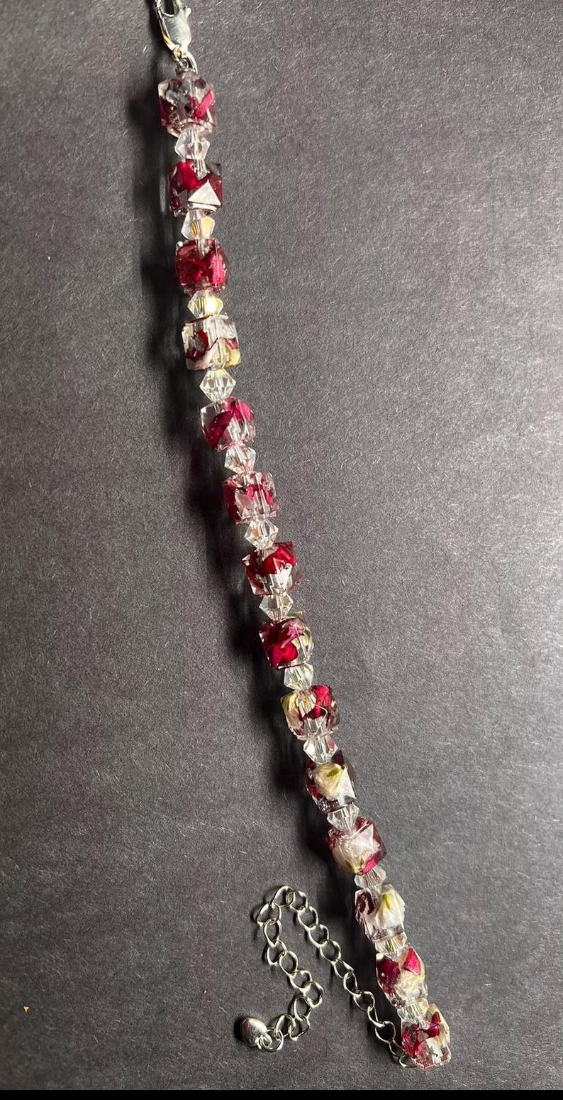 May include: A beaded bracelet with alternating clear and red square beads. The red beads have a floral design inside. The bracelet has a silver-colored clasp and chain extender.