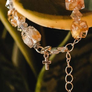 May include: A close-up of a bracelet featuring clear and amber-colored crystal beads. The bracelet has a silver-colored chain and a small cross charm. The background is a blurred green and yellow.