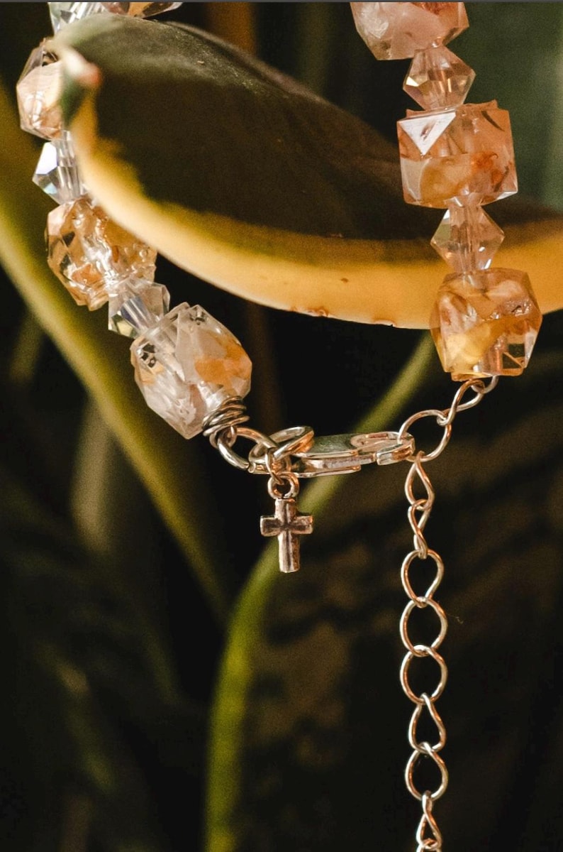 May include: A close-up of a bracelet featuring clear and amber-colored crystal beads. The bracelet has a silver-colored chain and a small cross charm. The background is a blurred green and yellow.