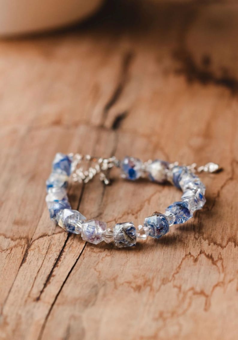 May include: A delicate bracelet featuring faceted, clear beads with blue and purple inclusions. The bracelet is strung on a silver chain and rests on a wooden surface, showcasing its intricate design and color.