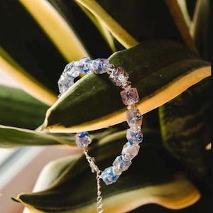 May include: A delicate bracelet featuring clear, square-cut beads with blue and purple accents. The bracelet is strung on a silver chain and is displayed against a backdrop of green and yellow leaves. The jewelry is a stylish accessory.