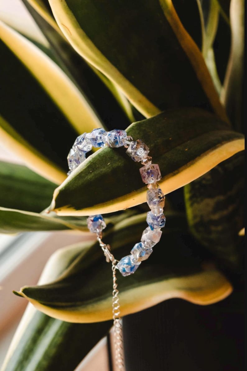 May include: A delicate bracelet featuring clear, square-cut beads with blue and purple accents. The bracelet is strung on a silver chain and is displayed against a backdrop of green and yellow leaves. The jewelry is a stylish accessory.