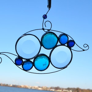 May include: A stained glass suncatcher with a whimsical design featuring blue and purple glass circles. The suncatcher is suspended from a black metal wire.