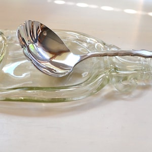 May include: A clear glass bottle, repurposed as a serving dish, with a silver ladle resting inside. The bottle has a unique, wavy shape.