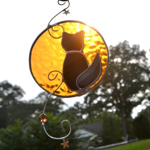 May include: A stained glass suncatcher featuring a black cat silhouette against a yellow sun. The cat is facing away from the viewer and has a long tail. The suncatcher is hanging from a metal hook and has a beaded chain.