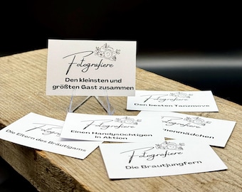 Könnte beinhalten: Eine Sammlung weißer Karten mit schwarzer Kalligraphie und floralen Illustrationen, angeordnet auf einer Holzoberfläche. Die Karten zeigen das Wort "Fotografiere" und verschiedene deutsche Sätze, wie "Den kleinsten und größten Gast zusammen."