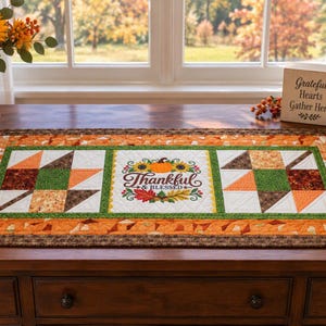 May include: A rectangular quilted table runner featuring a central panel with the text Thankful & Blessed surrounded by sunflowers and leaves, flanked by two leaf-patterned blocks in shades of orange, green, brown, and white.