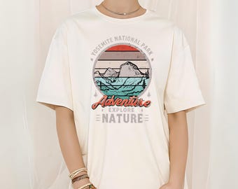 Könnte beinhalten: Elfenbeinfarbenes T-Shirt mit einem kreisförmigen Grafikdesign, das eine Berglandschaft, den Text "Yosemite National Park" und die Worte "Adventure Explore Nature" zeigt. Das Design enthält Rot-, Braun- und Blautöne. Das Shirt hat kurze Ärmel.