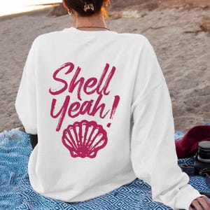 May include: White sweatshirt with the text "Shell Yeah!" in a magenta script font, along with a magenta seashell graphic. The sweatshirt is a casual style, suitable for beach or casual wear.