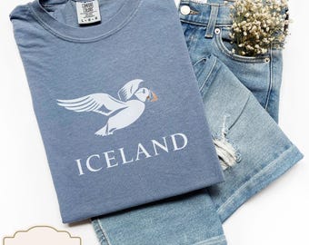 Op de afbeelding: Een blauw t-shirt met een witte papegaaiduiker afbeelding en het woord "ICELAND" gedrukt op de voorkant. Het t-shirt is gevouwen en gelegd op een blauwe spijkerbroek.