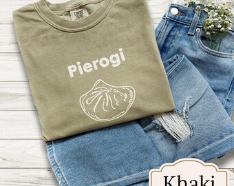 以下が含まれることがあります： 「Pierogi」の文字と白い餃子のグラフィックが入ったカーキ色のTシャツ。明るいブルーのデニムショーツと組み合わせています。「Khaki」の文字が小さなラベルに表示されています。