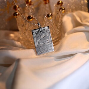 May include: A silver necklace with a pendant shaped like a trading card featuring a dragon illustration. The card has the text "3000 2500" on it.