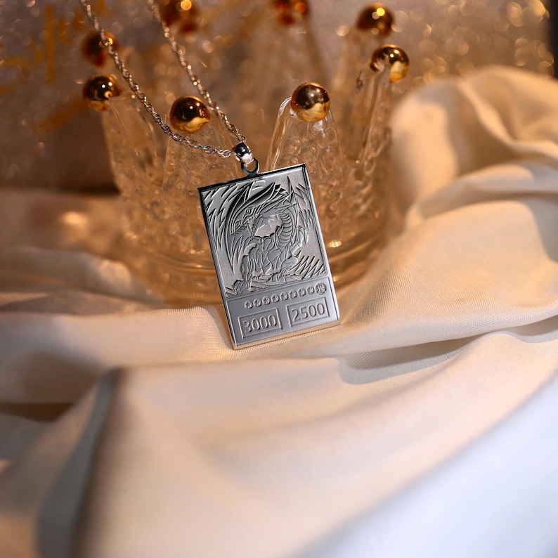 May include: A silver necklace with a pendant shaped like a trading card featuring a dragon illustration. The card has the text "3000 2500" on it.